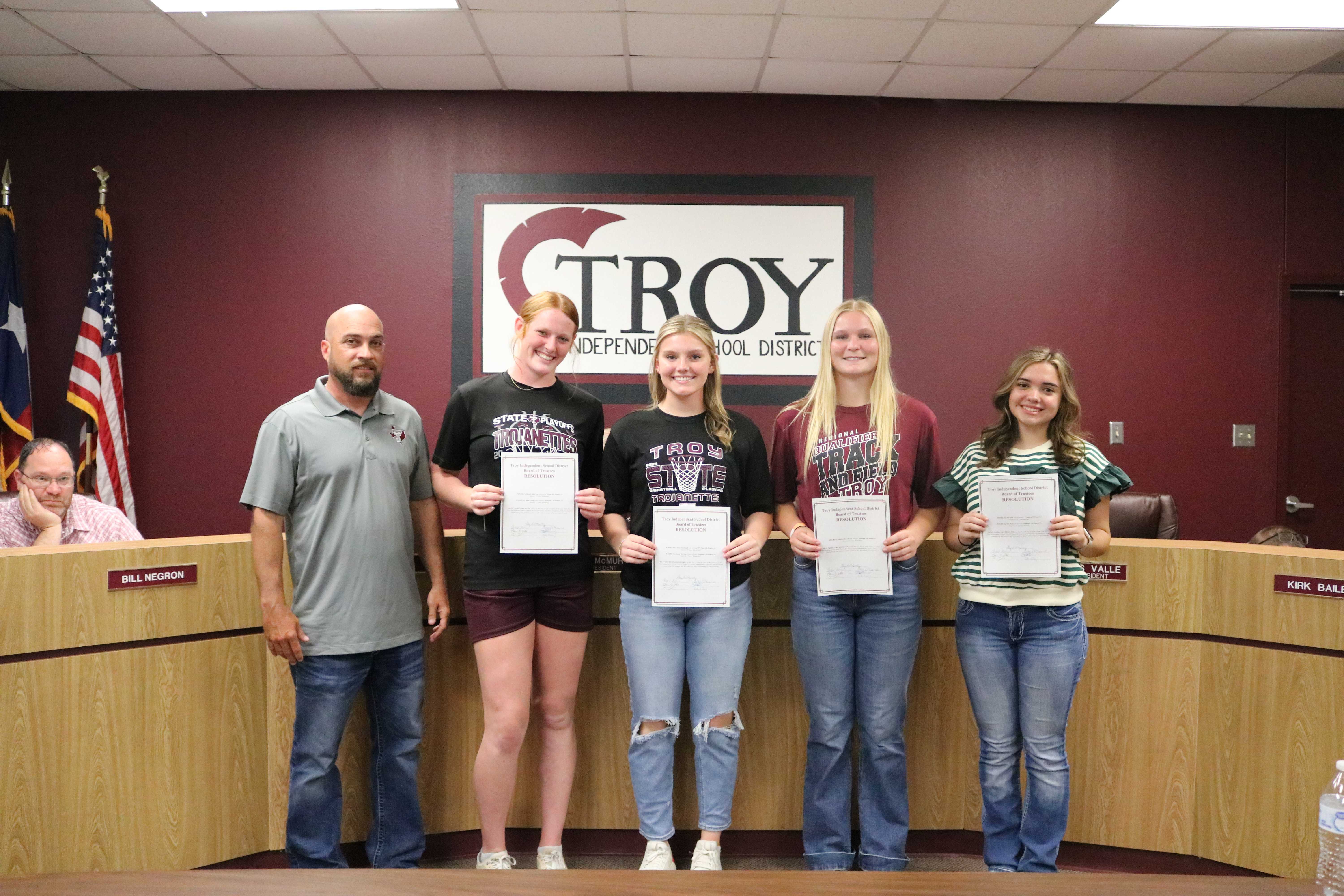 Four young women stand in front of a man, all holding certificates in a school board meeting room.
