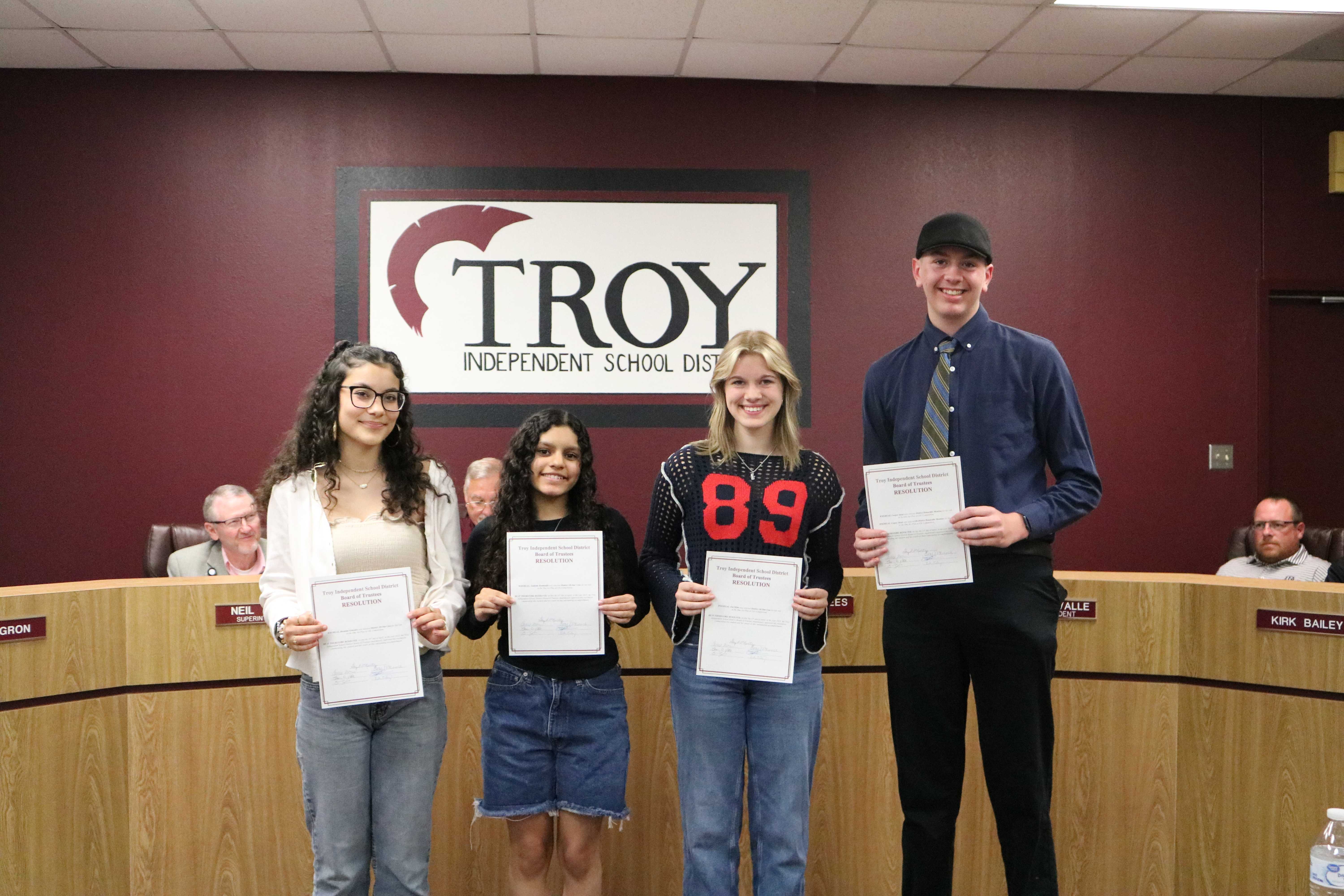 Four students stand in front of a school board, holding certificates of achievement.