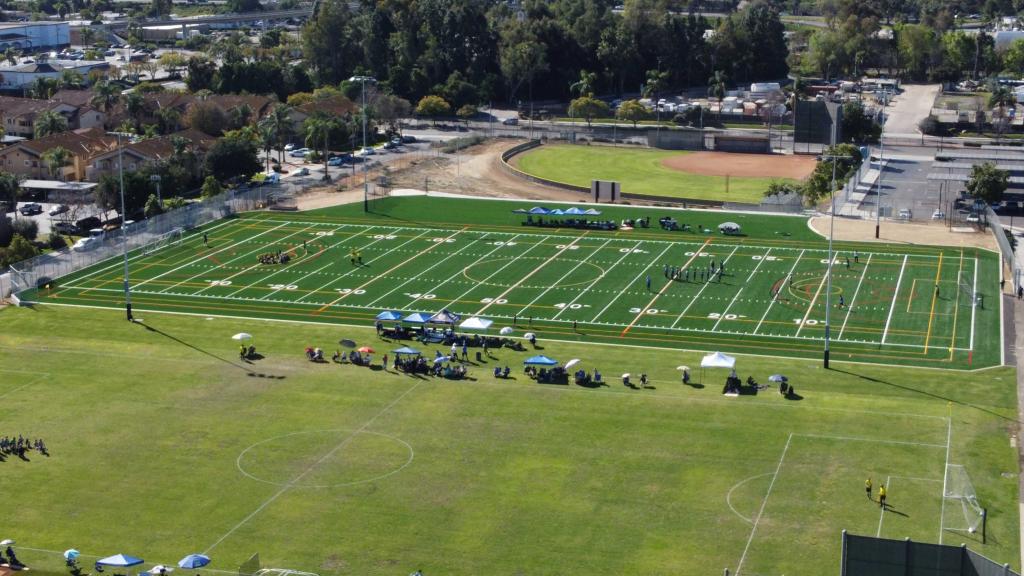 An aerial view shows a football field with players practicing and spectators gathered under tents.