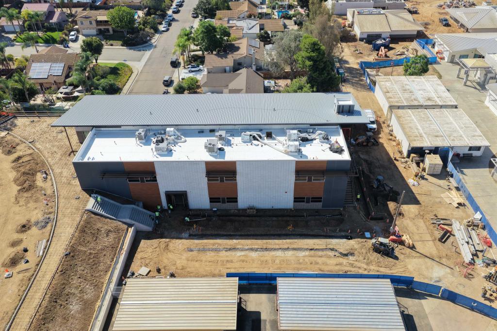 An aerial view of a modern building under construction, surrounded by dirt and neighboring houses.