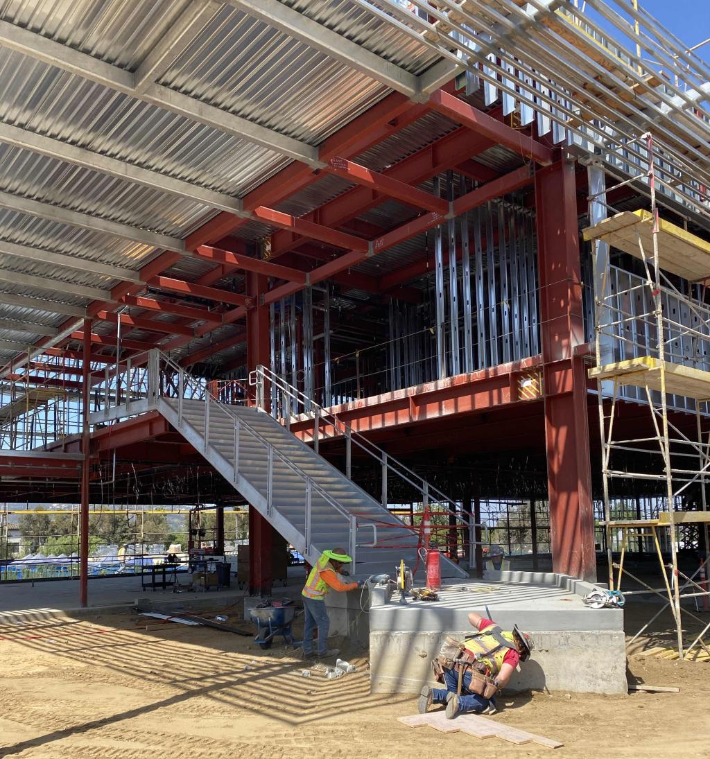 Construction workers in hard hats and safety vests work on a multi-level building with exposed steel beams and a metal roof.