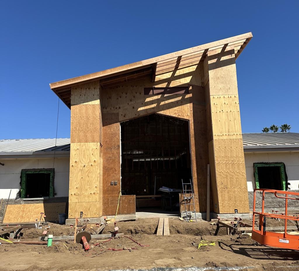A building under construction features a large wooden entryway with a slanted roof against a clear blue sky.