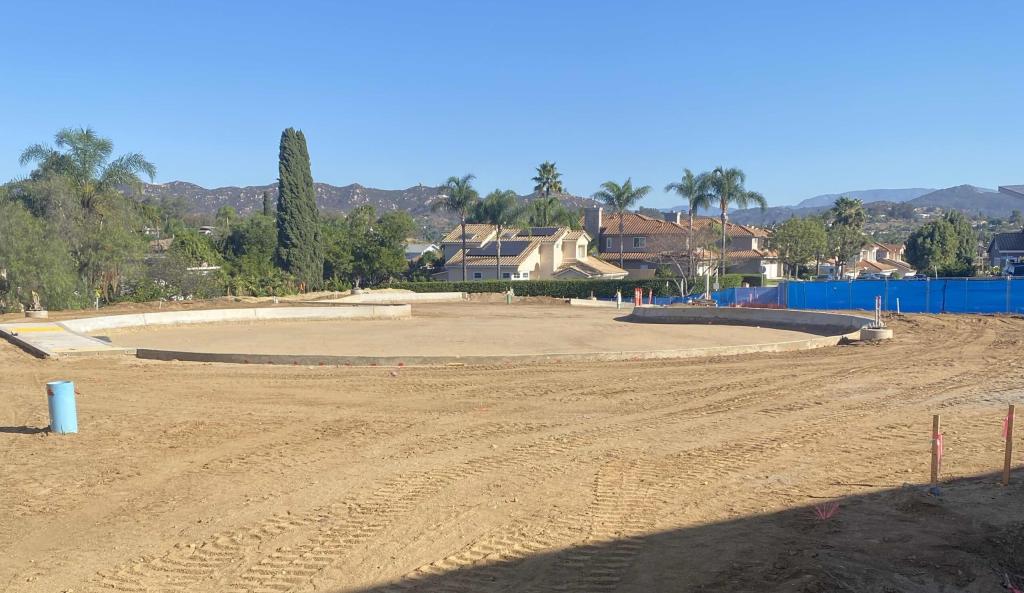 A wide dirt equestrian arena sits under a clear blue sky, surrounded by distant hills and trees.