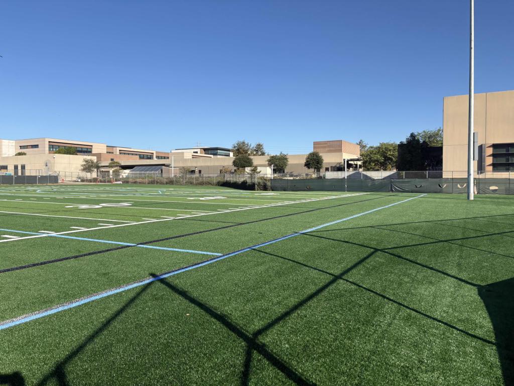 A wide-angle view of an empty green turf field under a clear blue sky.