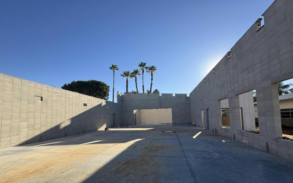 A wide-angle view of a building construction site under a clear blue sky, with palm trees visible in the background.