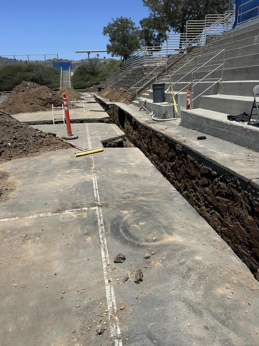 Construction site with a deep trench dug in concrete, alongside stadium seating.