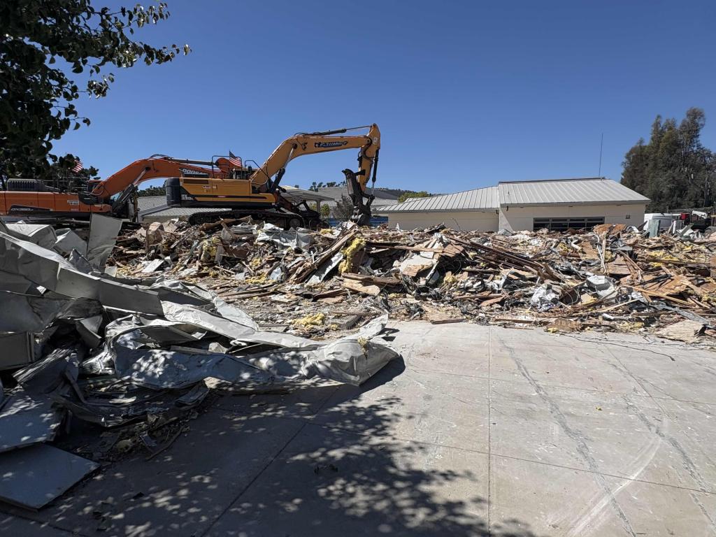 An excavator works on a pile of rubble from a demolished building under a clear blue sky.