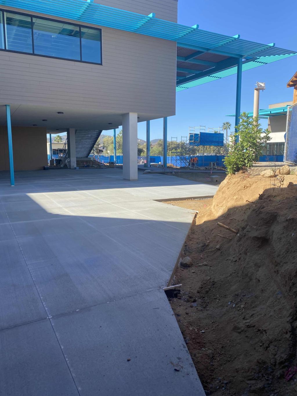 A modern building under construction features a newly paved concrete courtyard and a blue awning against a bright sky.