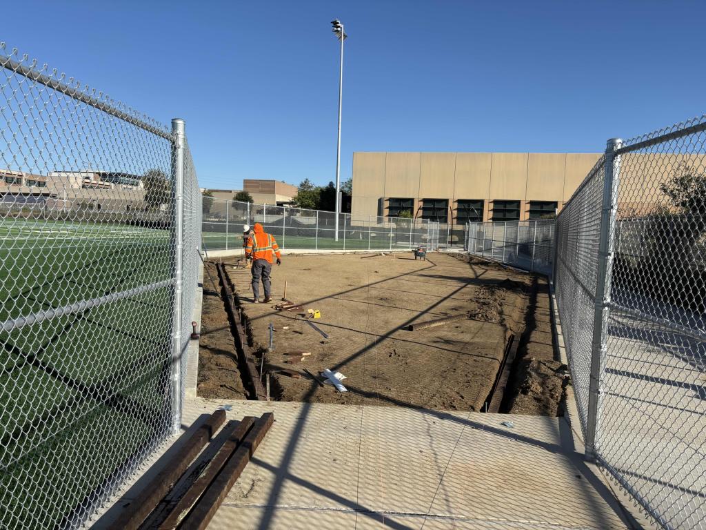 A worker in an orange vest stands near a trench excavation between chain-link fences under a clear blue sky.