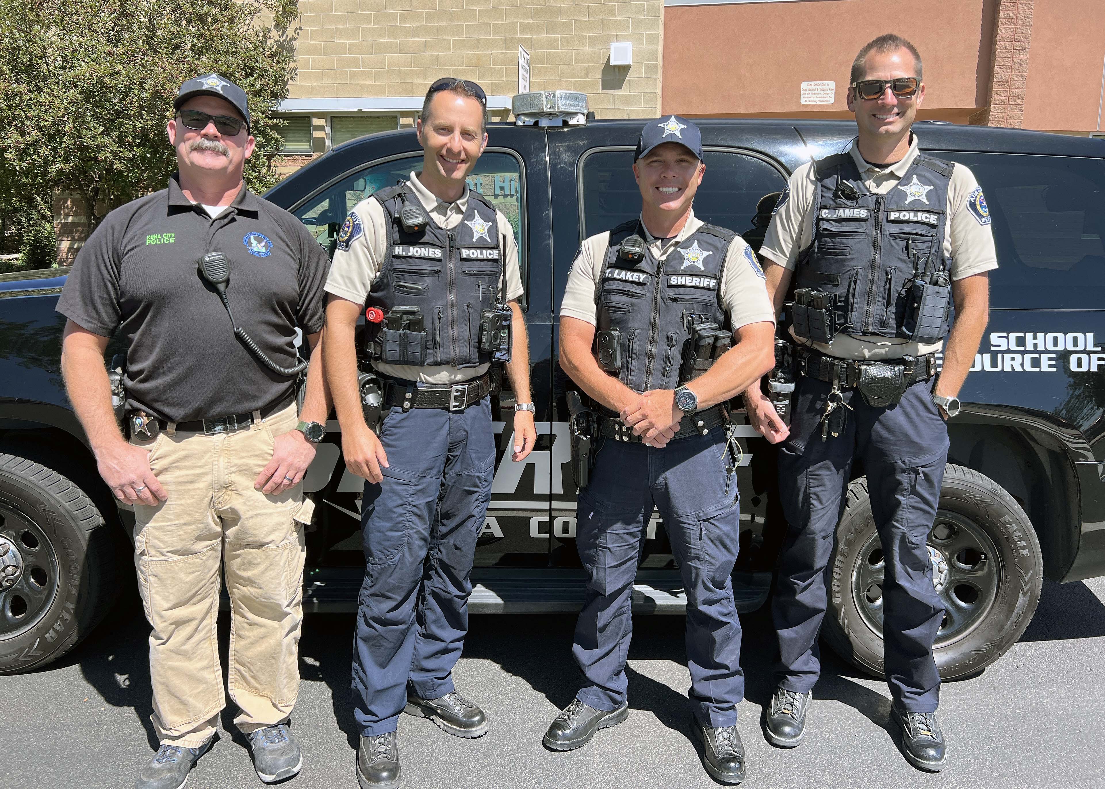 Four law enforcement officers stand in front of a black vehicle, smiling at the camera.