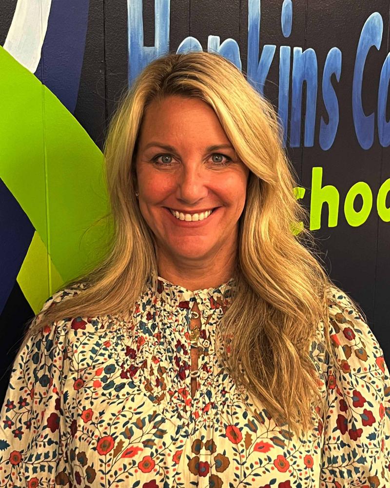 A smiling woman with blonde hair, wearing a floral shirt, stands in front of a colorful background.