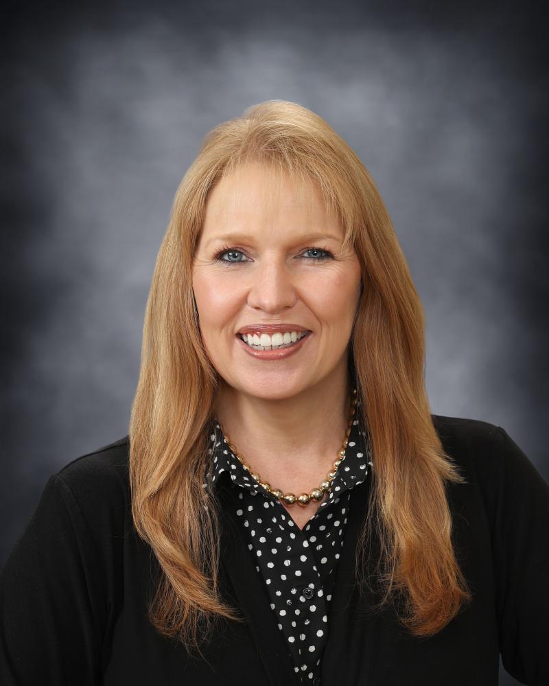 A professional headshot of a smiling woman with long blonde hair, wearing a black top and polka-dot shirt.