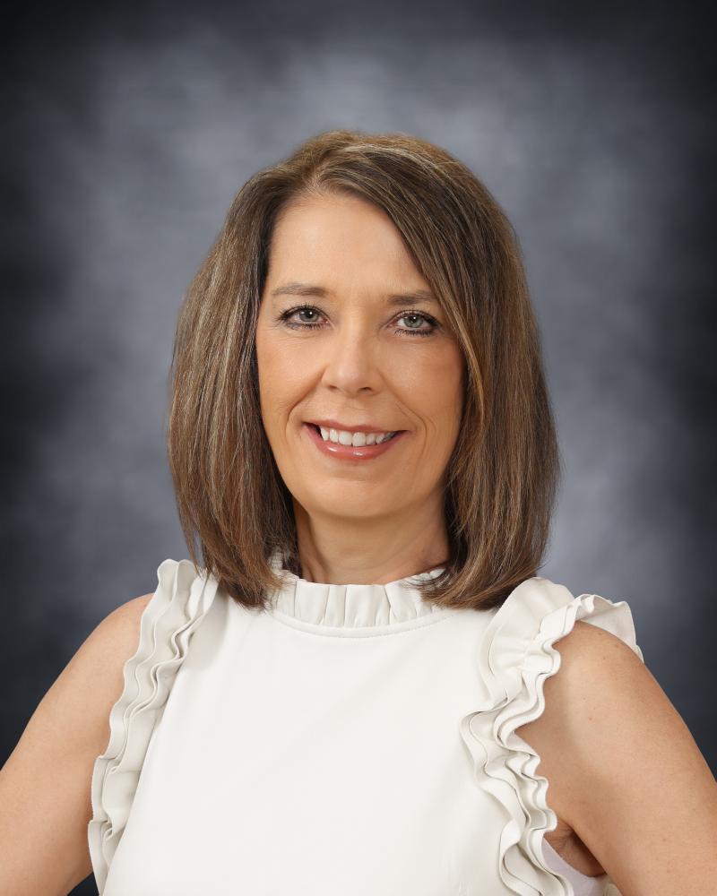 A professional headshot of a smiling woman with shoulder-length brown hair, wearing a white sleeveless top with ruffled details.