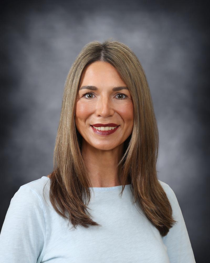 A professional headshot of a woman with long brown hair, smiling gently against a dark, mottled background.