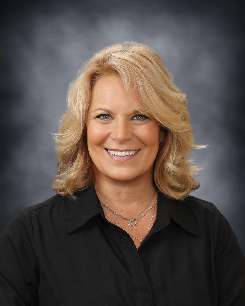 A professional headshot of a smiling woman with blonde hair, wearing a black shirt against a dark, textured background.