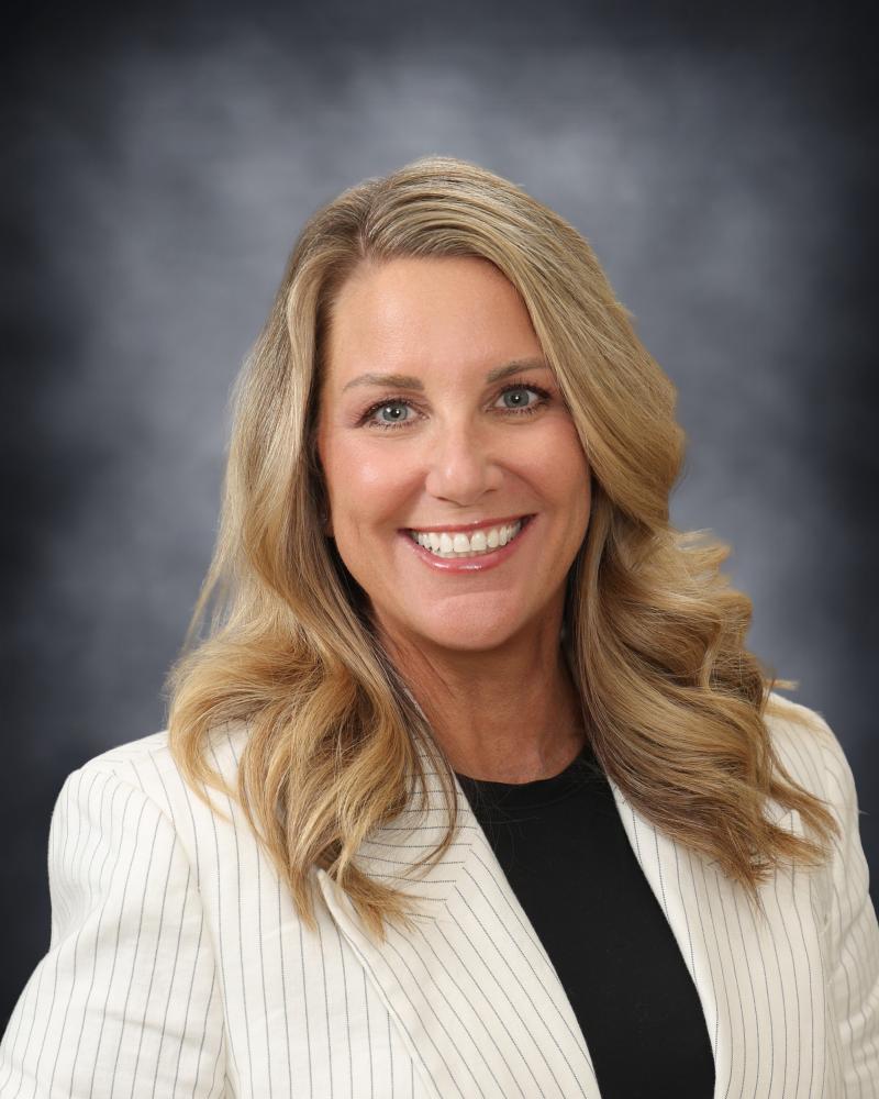 A professional headshot of a smiling woman with long blonde hair, wearing a white blazer over a black top against a dark, mottled background.