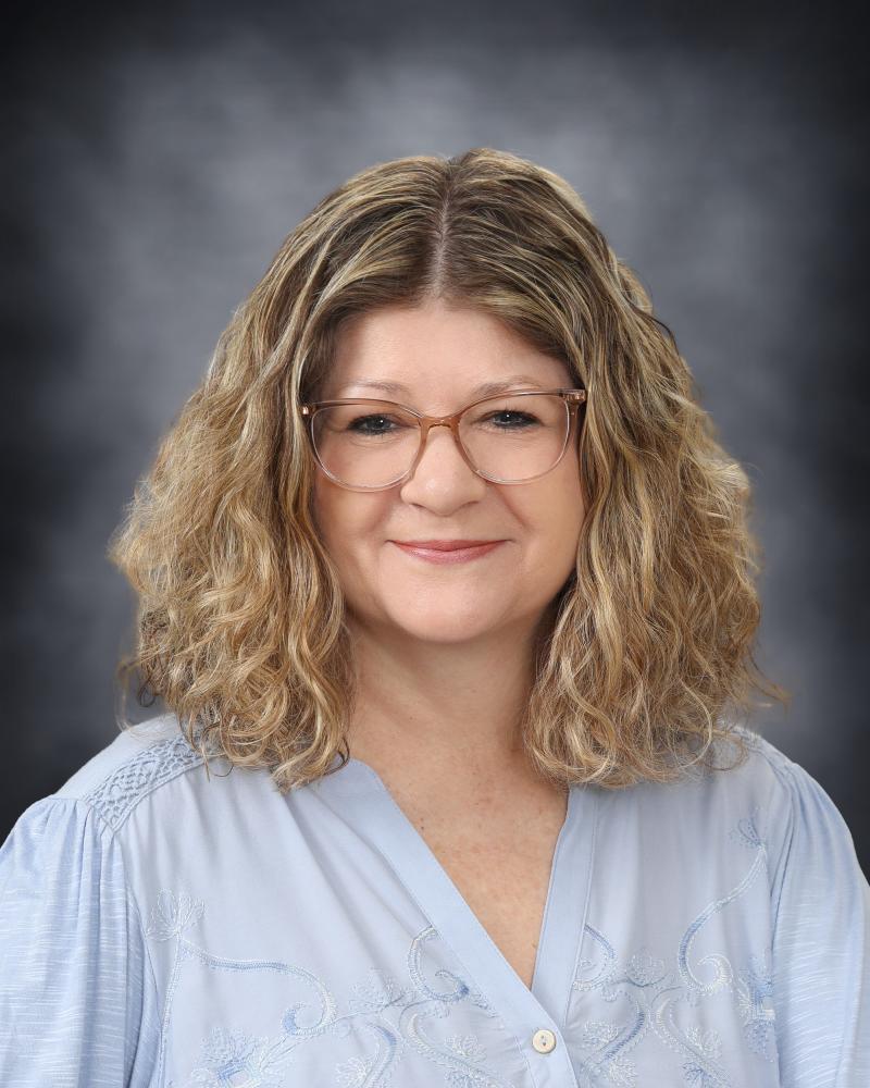 A portrait of a woman with shoulder-length, curly blonde hair and glasses, wearing a light blue top against a dark, mottled background.