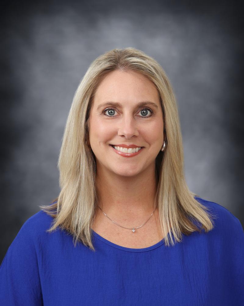 A professional headshot of a smiling woman with blonde hair, wearing a bright blue shirt against a gray background.