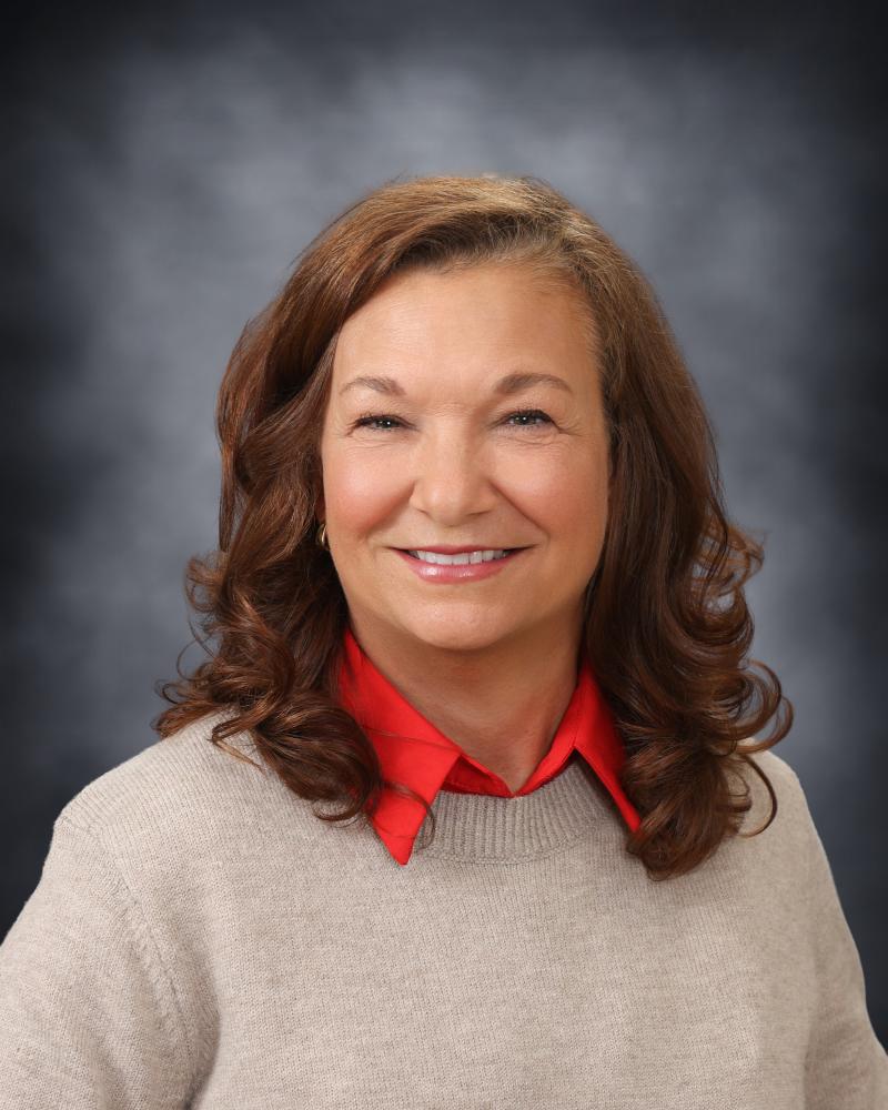 A portrait of a smiling woman with shoulder-length brown hair, wearing a red shirt under a beige sweater.
