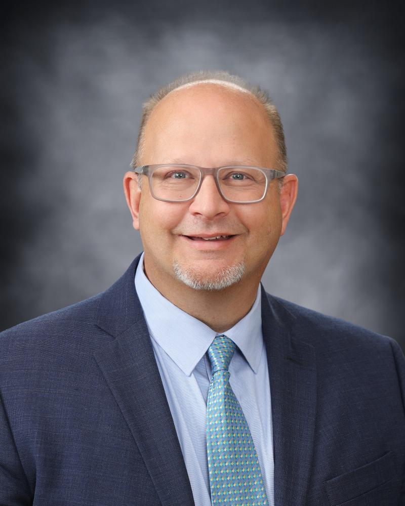 A portrait of a smiling man wearing glasses, a blue suit jacket, a light blue dress shirt, and a teal patterned tie against a mottled grey background.
