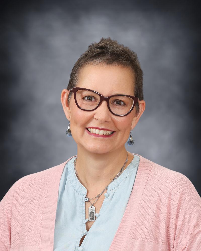 A portrait of a smiling woman wearing glasses and a pink cardigan against a gray background.