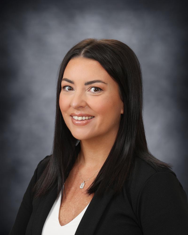 A professional headshot of a smiling woman with dark hair wearing a black blazer and white top against a grey textured background.