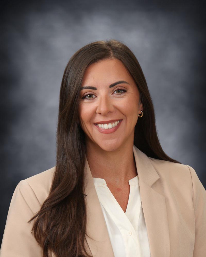 A professional headshot of a smiling woman with long brown hair, wearing a beige blazer over a white top.