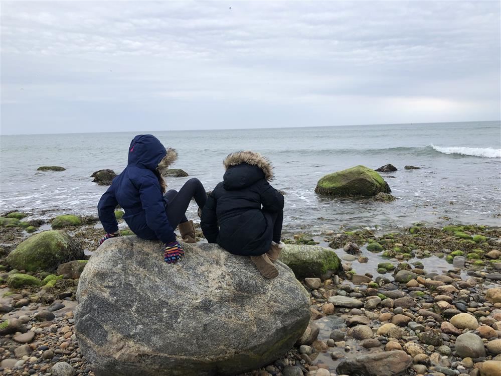 Two kids sitting on a beach rock, looking out at the sea under the clear blue sky.