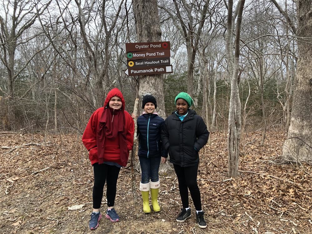 Three girls smiling in front of a wooden sign amidst a lush forest