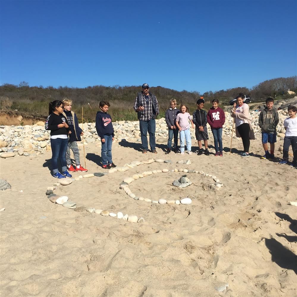 A circle formed by people on the beach, with the ocean waves gently crashing in the background.