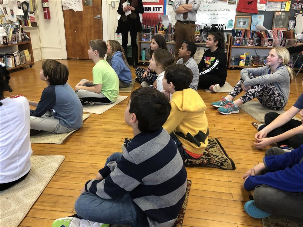 Students sitting on the floor in a classroom, attentively listening to their teacher's instructions.
