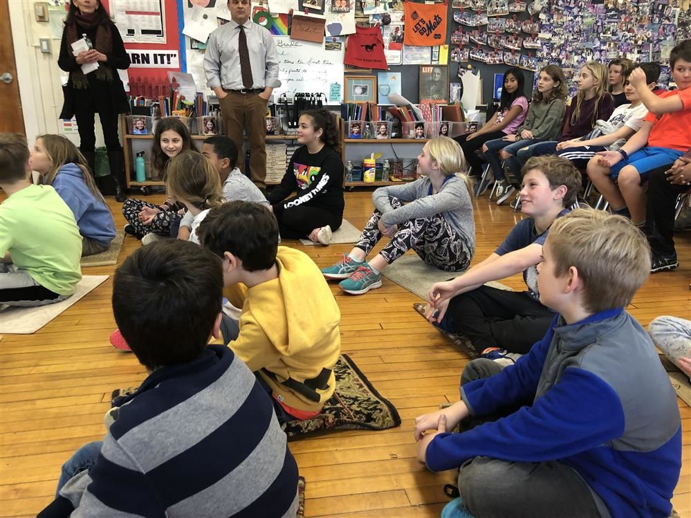 Students sitting on classroom floor listening to teacher.
