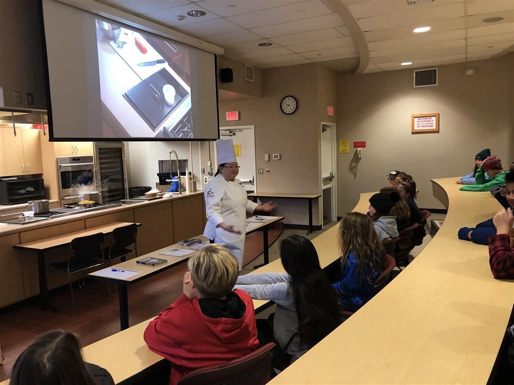 A woman in a white coat giving a presentation to a class of students.