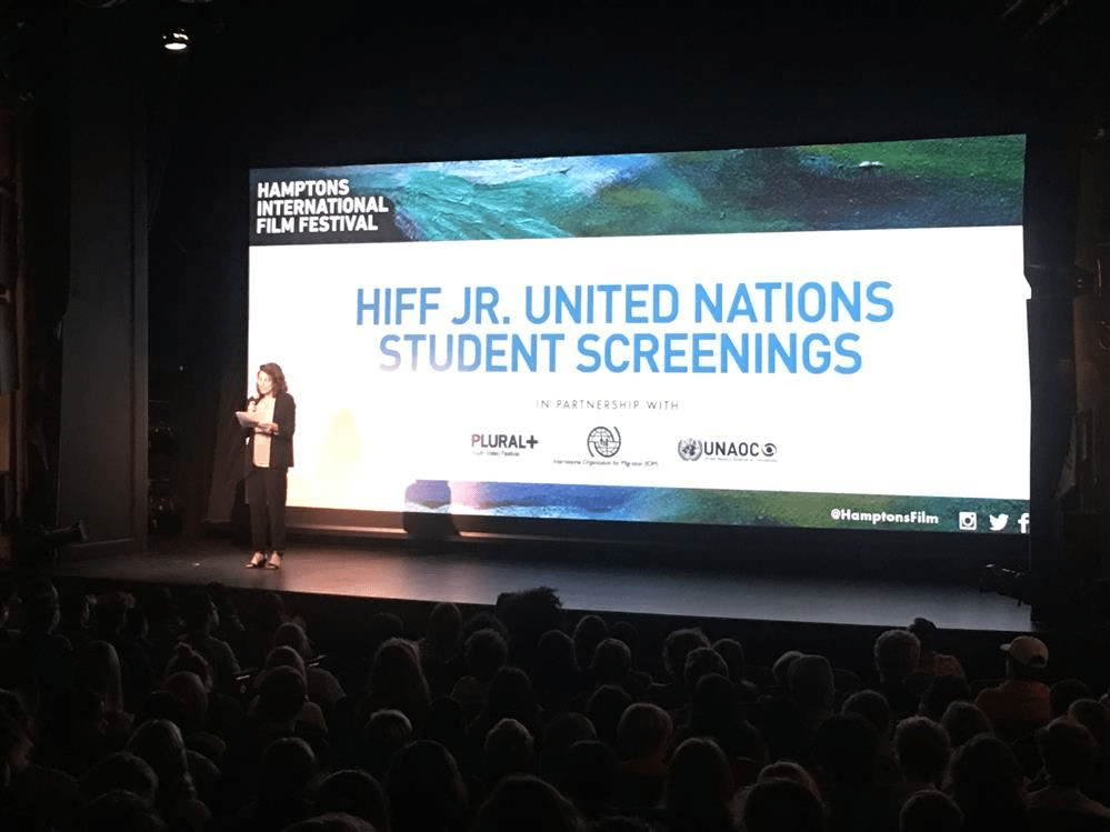 A woman presenting on stage at a conference with a large screen displaying a presentation.