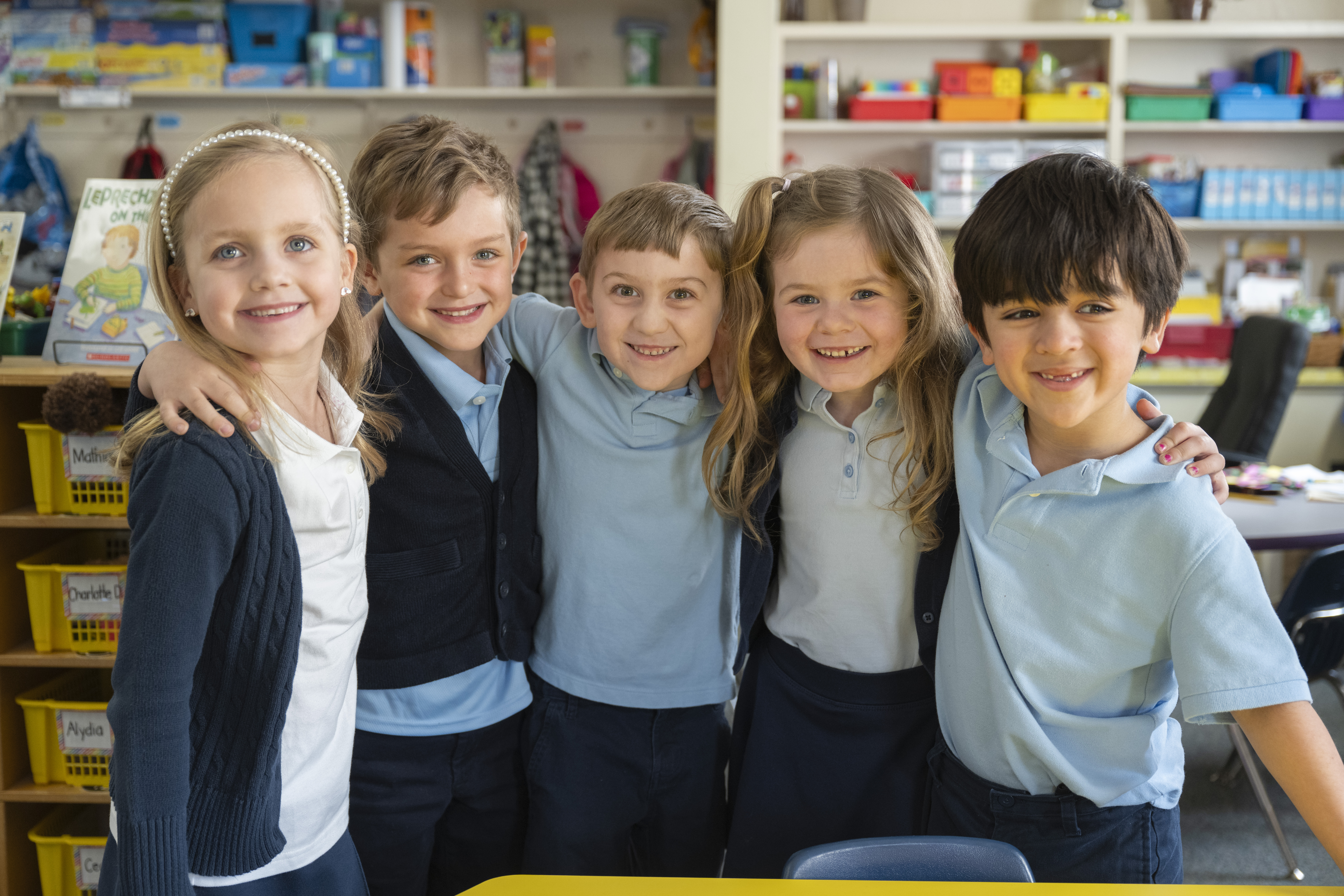 Five smiling children stand together, arms around each other, in a classroom.