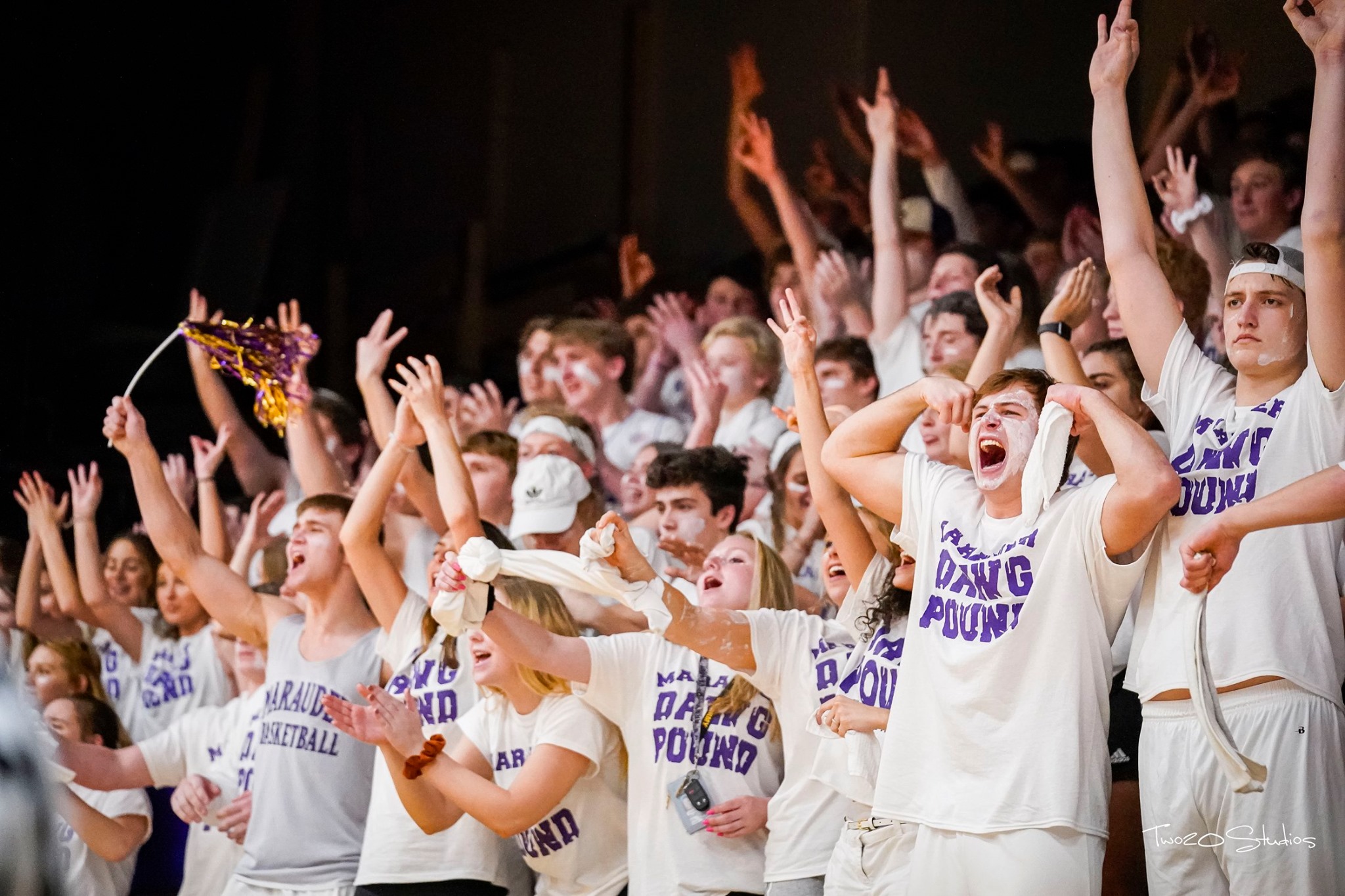 A crowd of people in white shirts cheer with their arms raised.