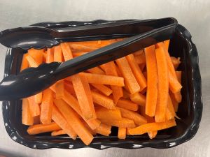 Freshly cut carrot sticks are displayed in a black container with tongs.