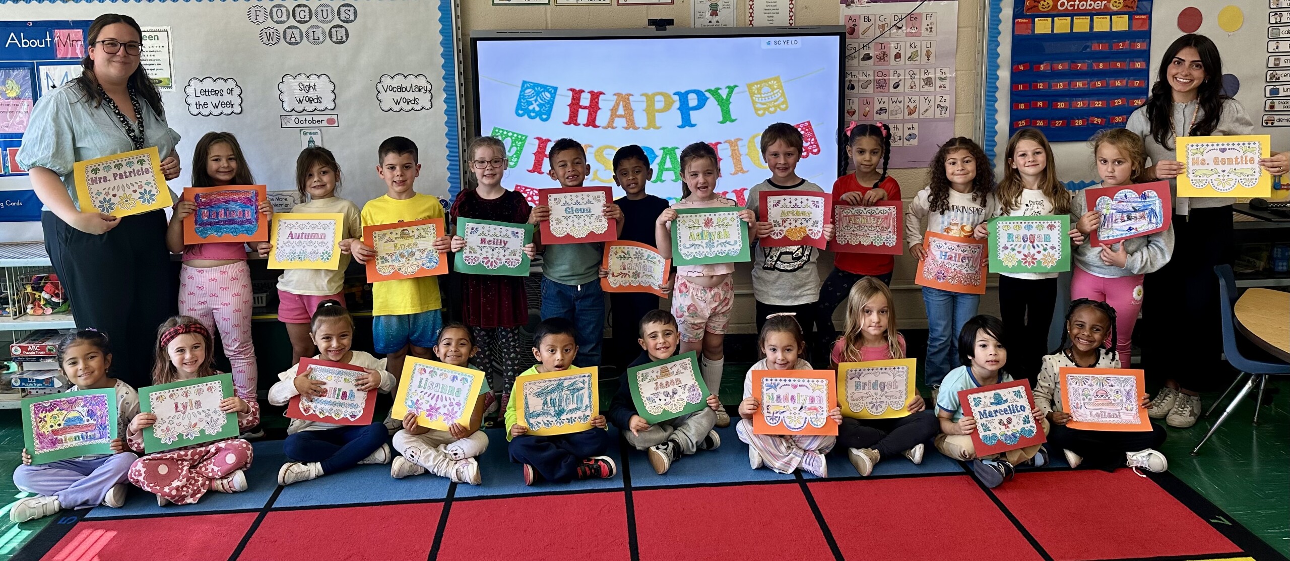 A group of students and two teachers pose with certificates in a colorful classroom.