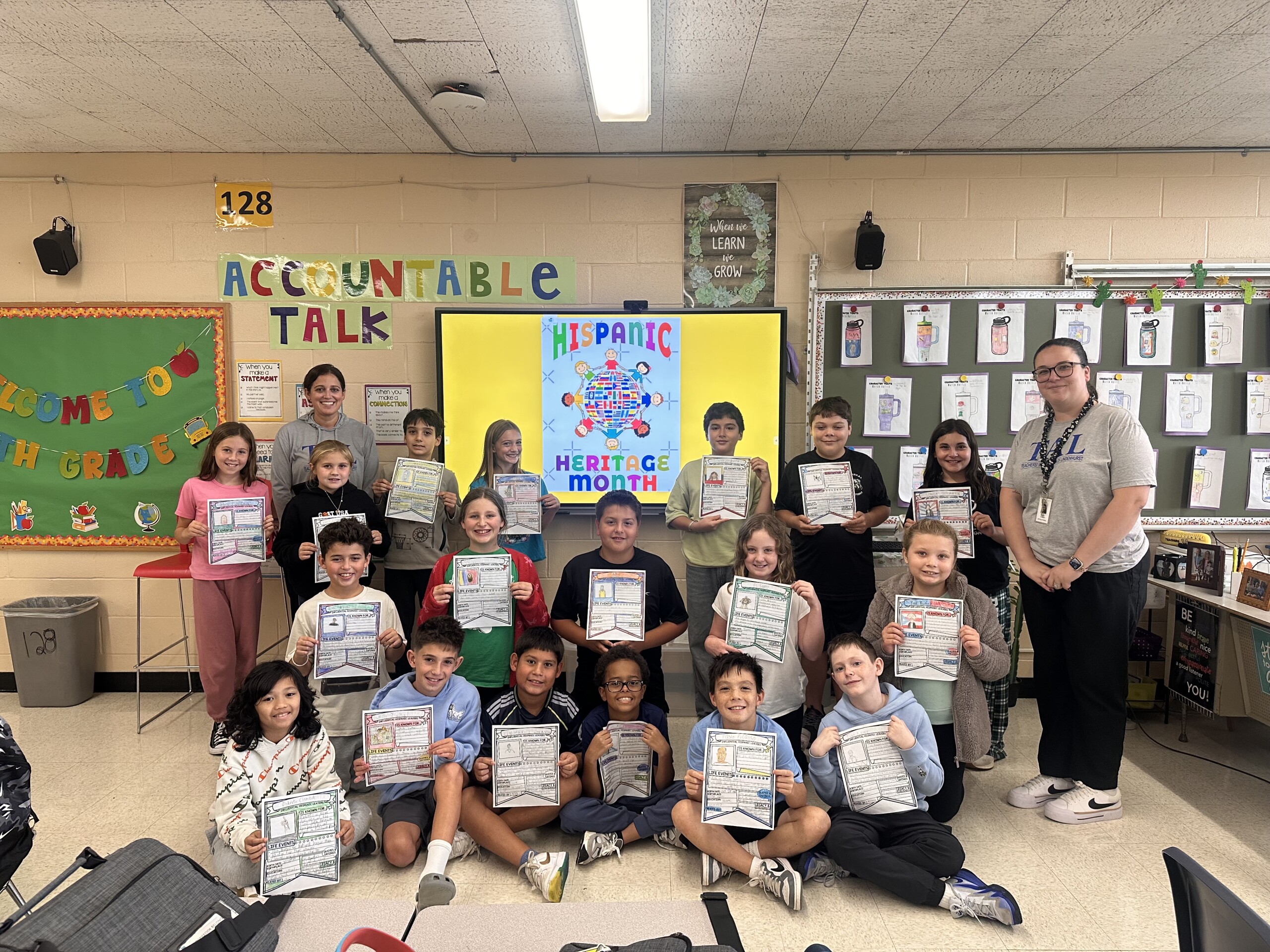 A teacher and students pose together in a brightly lit classroom, holding up their work.