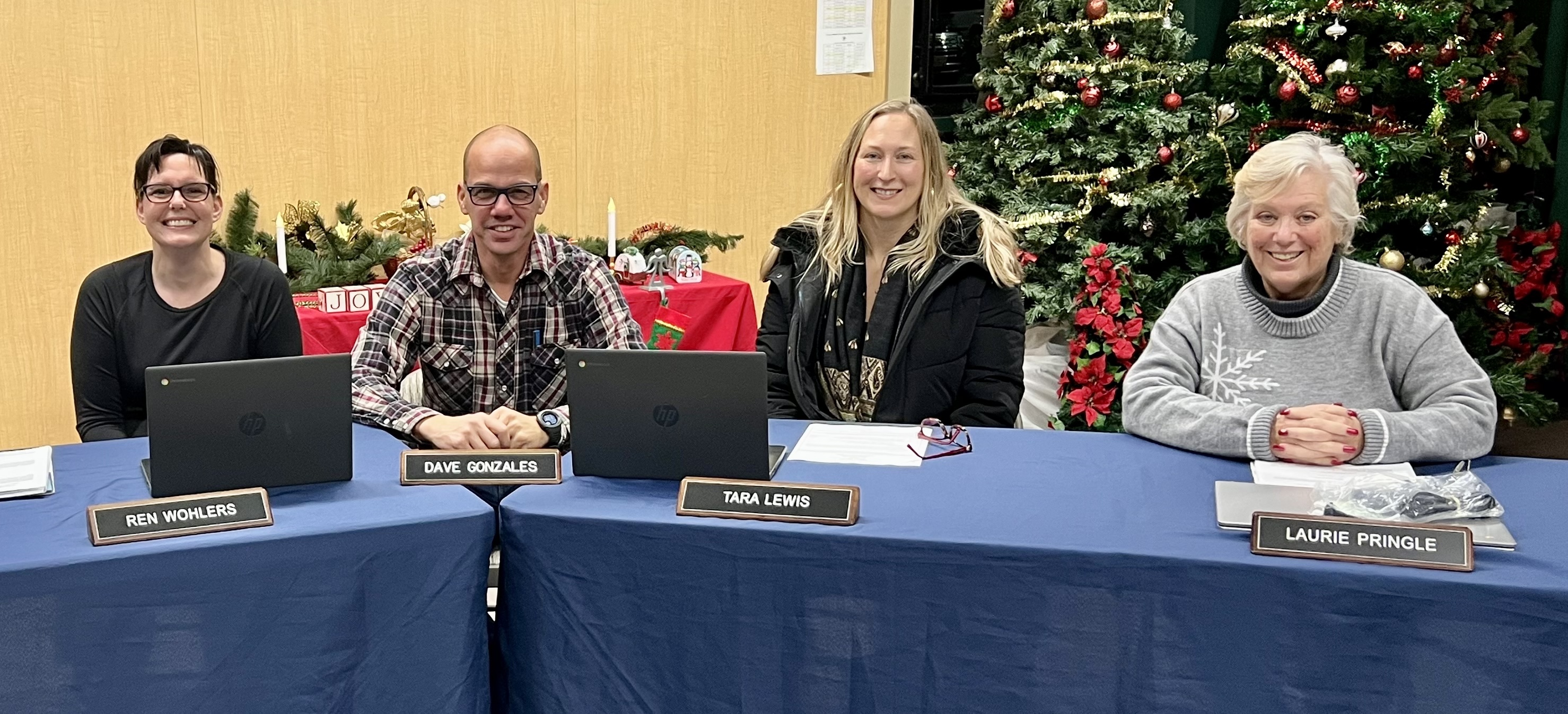 Four people sit at a table with laptops, in front of a Christmas tree.