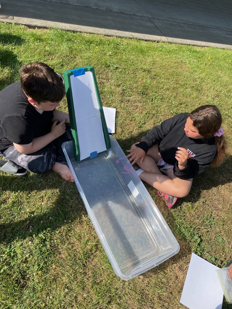 Two students work together on a science experiment outdoors on the grass.