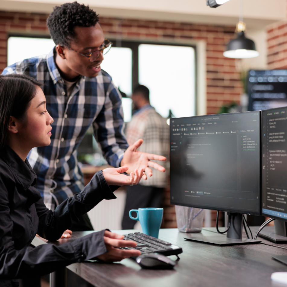 Two colleagues collaborate at a desk in a bright office, reviewing code on dual monitors.
