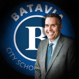 A smiling man in a suit and tie stands in front of a blue Batavia City Schools logo.