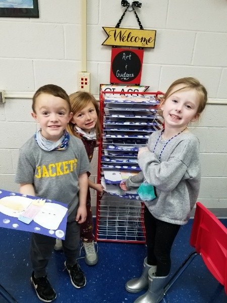 A group of children are standing in front of a bulletin board. They are holding up their artwork.