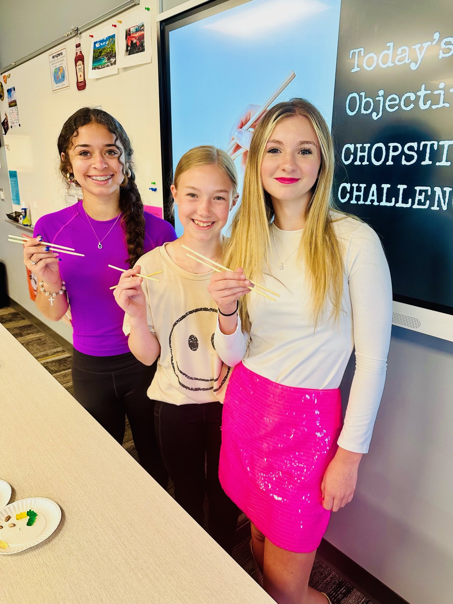 Three students are standing in front of a board. They are holding chopsticks.