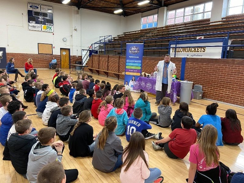 A group of students are sitting on the floor in a school gym. They are listening to a man in a lab coat who is standing in front of them.