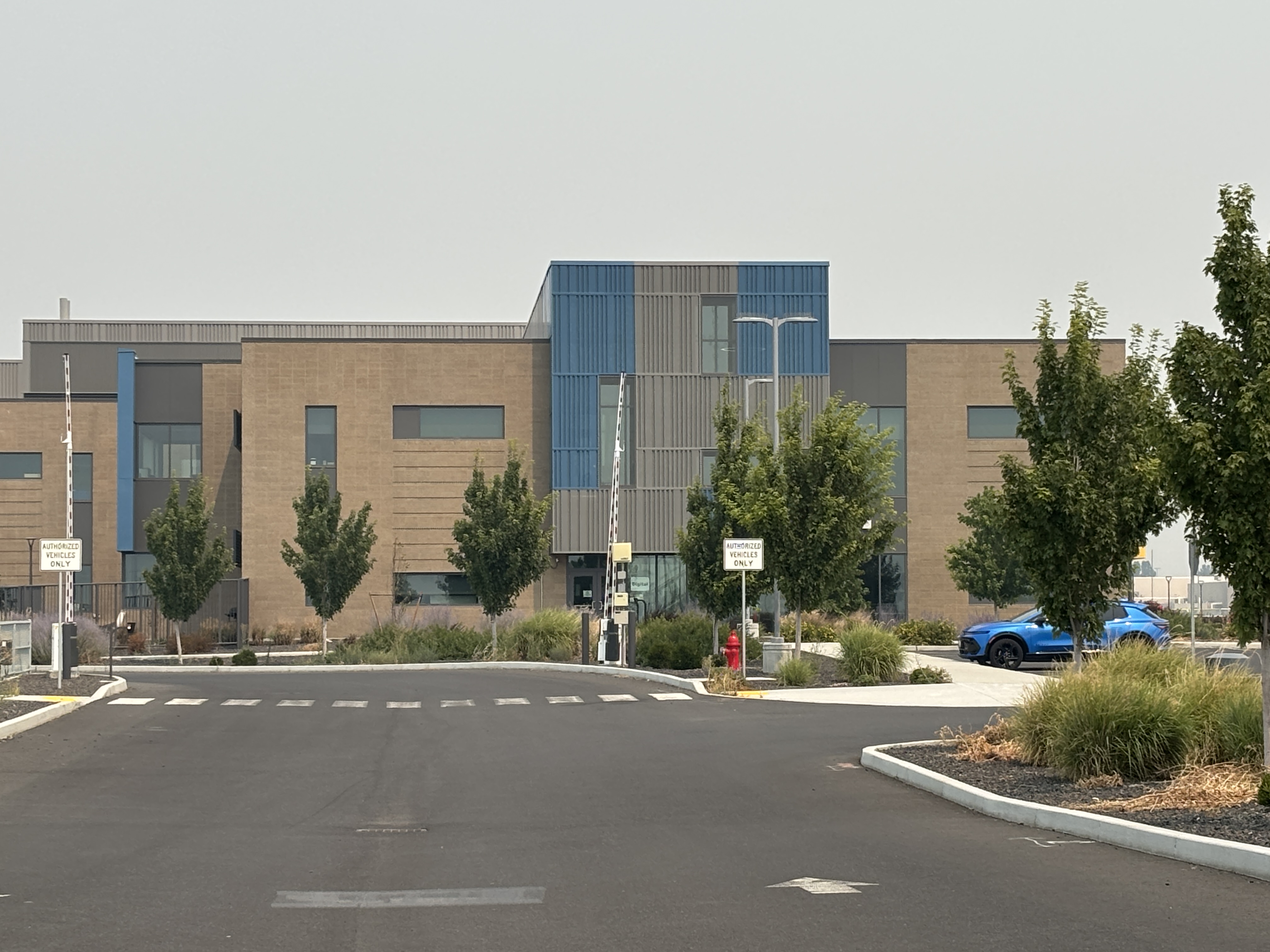 A modern building with tan and blue facade, featuring trees and two blue cars parked outside.