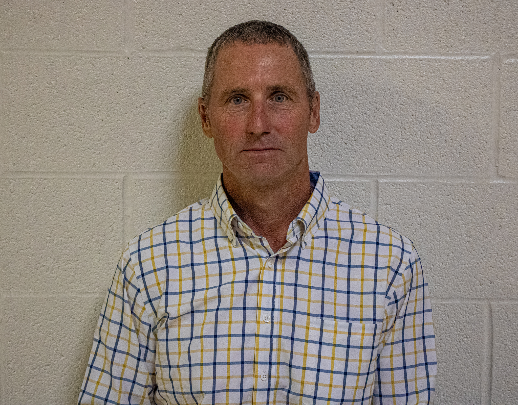 A middle-aged man with short, graying hair stands against a light-colored brick wall.
