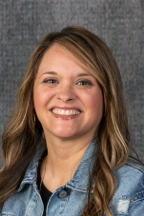 A headshot of a woman with long brown hair. She is wearing a blue jean jacket and smiling.