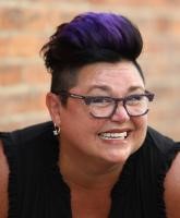 A woman with purple hair and glasses smiles warmly at the camera.